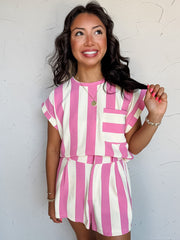 A three-quarter length photograph of a smiling woman with dark wavy hair touching a strand of her hair while posing against a textured white wall. She is wearing a short-sleeved top made from a 58% polyester, 30% cotton, 10% rayon, and 2% spandex blend, featuring wide pink and off-white vertical stripes, a chest pocket, and cuffed cap sleeves, paired with matching striped shorts and styled with a round pendant necklace and gold beaded bracelets.