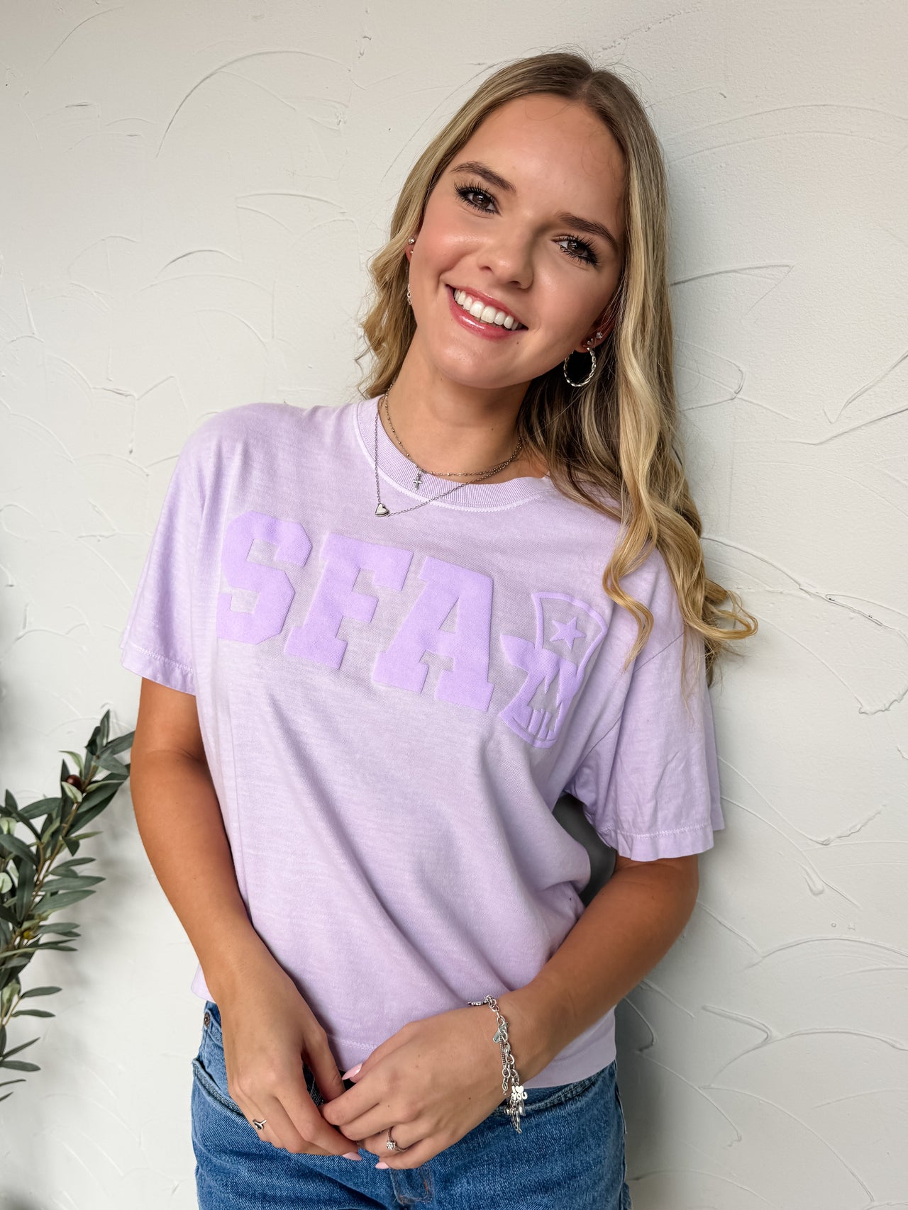 Woman wearing a light purple t-shirt with 'SFA' text and denim shorts against a white textured wall.