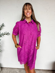 A three-quarter length photograph of a smiling woman with straight brown hair and bangs posing with her hands in her pockets against a textured white wall. She is wearing a vibrant magenta purple short-sleeved mini shirt dress made from 100% cotton, featuring all-over floral eyelet cutouts, a pointed shirt collar, a button-down front, puff sleeves, and a scalloped eyelet hem, styled with a colorful beaded necklace, stacked gold bangles, and delicate gold chain bracelets.