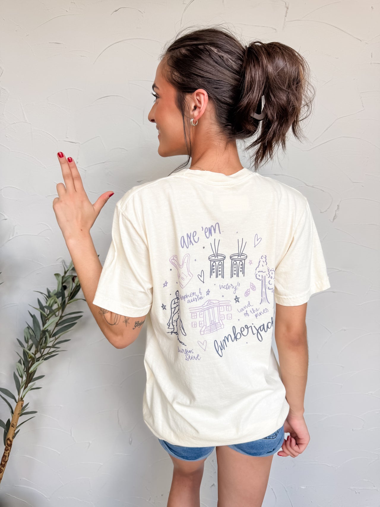 Woman wearing a white t-shirt with text and graphics on the back, standing against a plain wall.