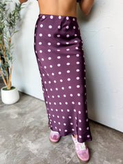 A waist-down photograph of a woman posing against a textured white wall next to a potted olive plant. She is wearing a high-waisted plum purple satin midi skirt made from 100% polyester, featuring a light pink polka dot pattern throughout and a fluid bias-cut fit, paired with retro pink suede sneakers with white laces.