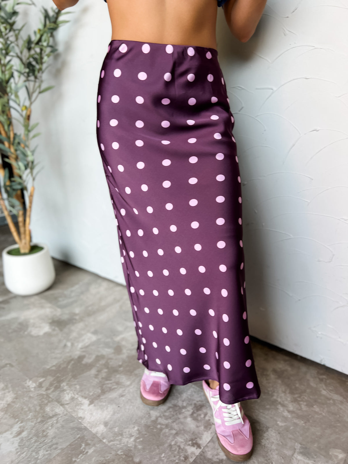A waist-down photograph of a woman posing against a textured white wall next to a potted olive plant. She is wearing a high-waisted plum purple satin midi skirt made from 100% polyester, featuring a light pink polka dot pattern throughout and a fluid bias-cut fit, paired with retro pink suede sneakers with white laces.