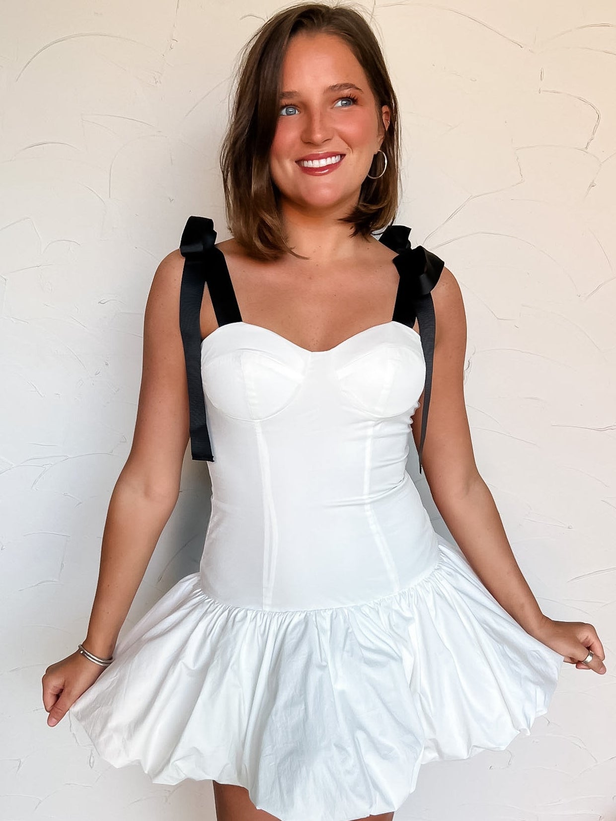 Woman wearing a white dress with black bow straps and a bubble hem skirt against a plain background