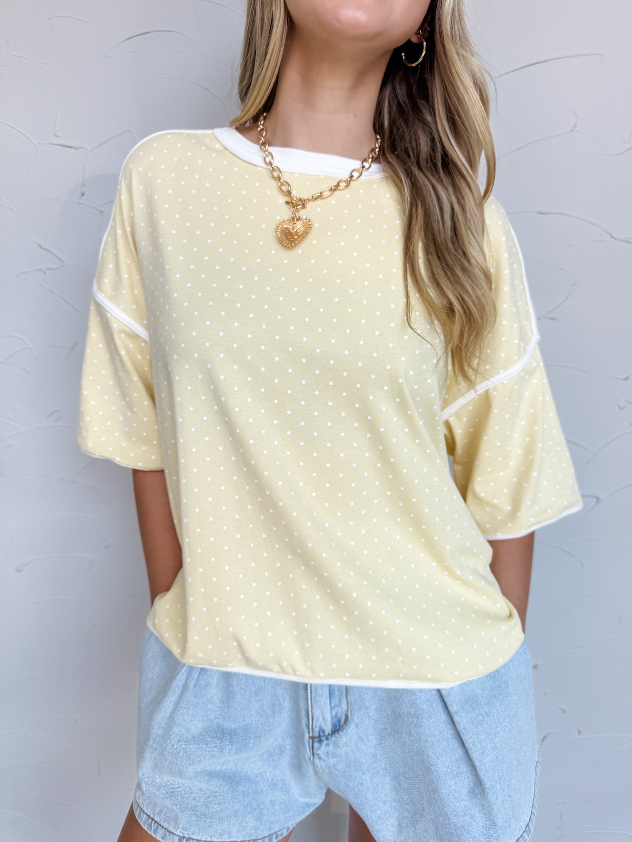 Smiling model wearing a relaxed-fit Yellow top made from a soft cotton-spandex knit fabric. The top features an allover white polka dot print, crew neckline, dropped shoulders, short sleeves, and white contrast binding at the neckline, sleeve cuffs, and hemline. The lightweight fabric has a comfortable feel with slight stretch. Styled with distressed light wash denim shorts, layered gold necklaces, gold hoop earrings, and loose wavy hair. Photo is taken against a neutral studio background with the model