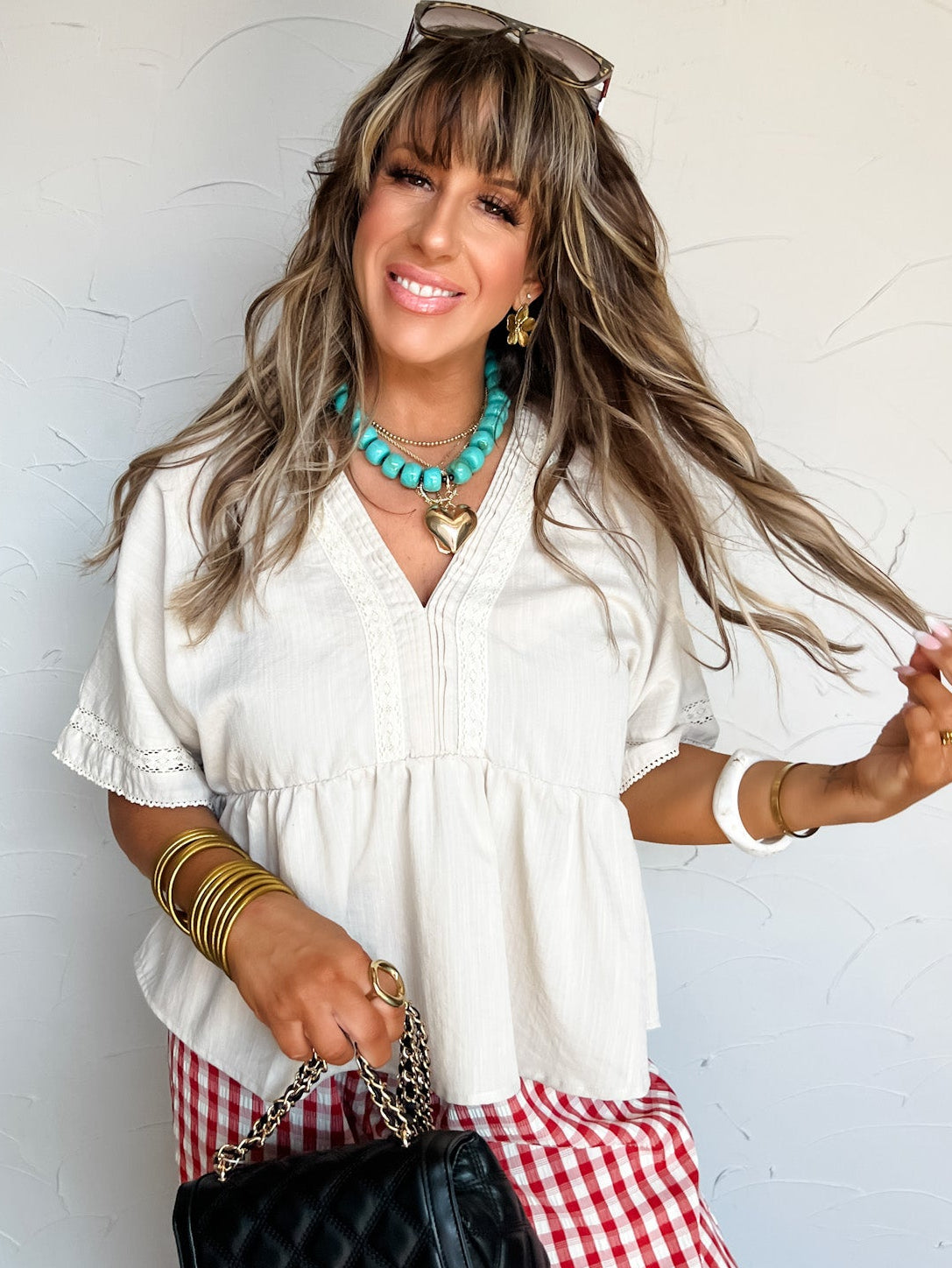 Woman in a white blouse and red checkered pants holding a black handbag against a white wall.