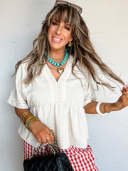 Woman in a white blouse and red checkered pants holding a black handbag against a white wall.