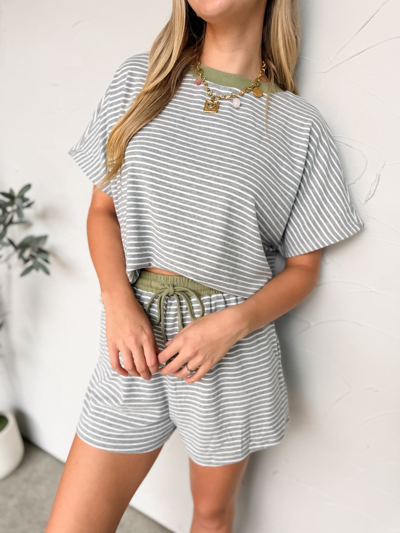 A three-quarter length photo of a smiling blonde woman looking off-camera against a textured white wall. She is wearing a matching grey and white fine-striped lounge set featuring a short-sleeved top with an olive green crew neckline. She playfully lifts the front of her loose shirt with both hands to showcase the contrasting olive green elastic waistband and functional drawstring tie of the matching high-waisted shorts. 