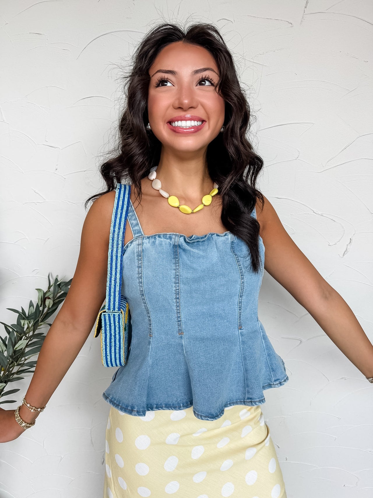 A smiling woman with dark wavy hair stands against a textured white wall, wearing a medium-wash denim peplum top. The tank-style blouse is crafted from a stretch denim blend of 72% cotton, 24% polyester, 3% rayon, and 1% spandex, featuring topstitched corset-style paneling, thick shoulder straps, and a flared hemline. She is styled with a yellow and white polka dot skirt, a chunky yellow and white stone necklace, stacked gold bracelets, and a blue striped beaded shoulder bag.