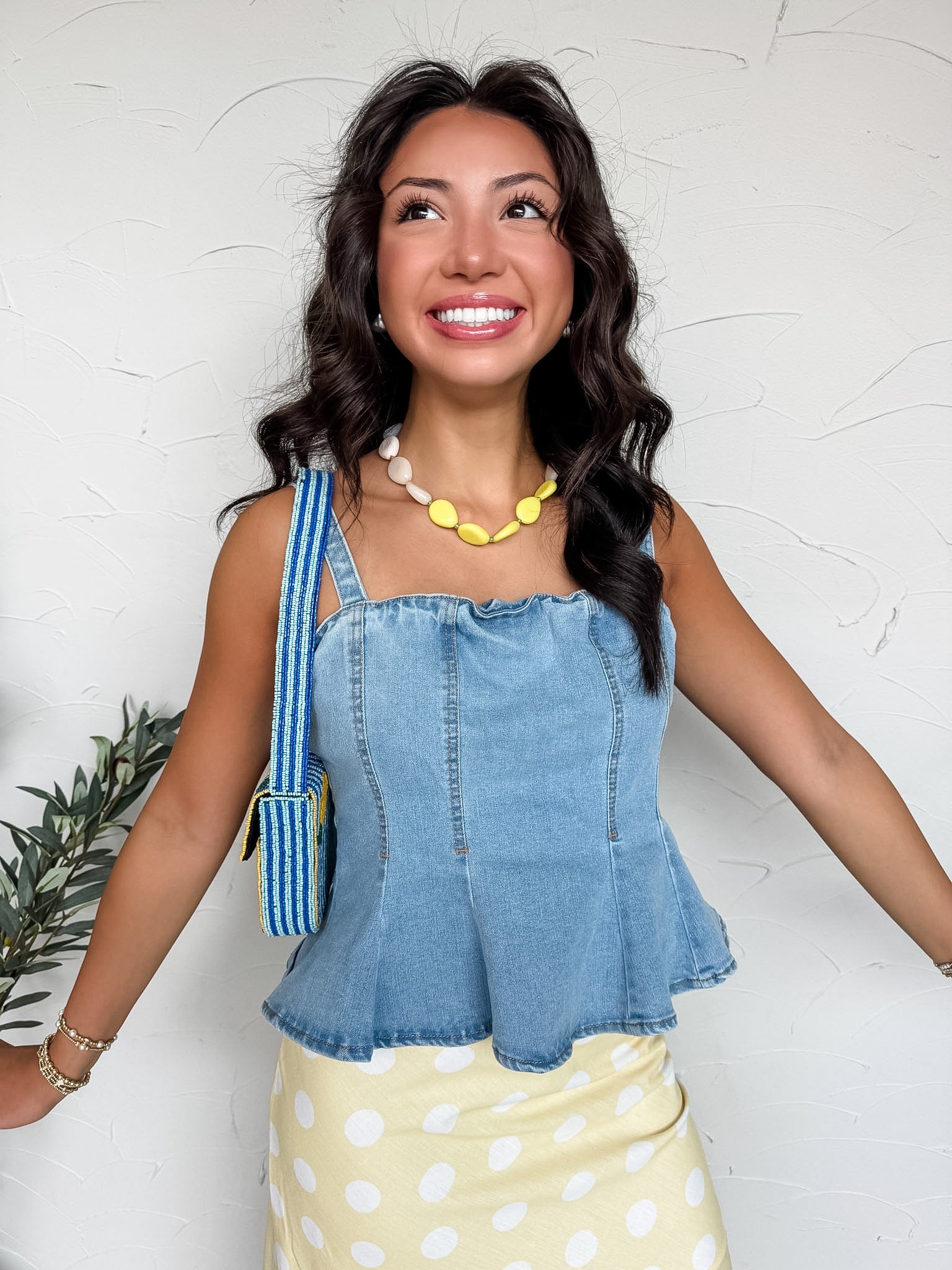 A smiling woman with dark wavy hair stands against a textured white wall, wearing a medium-wash denim peplum top. The tank-style blouse is crafted from a stretch denim blend of 72% cotton, 24% polyester, 3% rayon, and 1% spandex, featuring topstitched corset-style paneling, thick shoulder straps, and a flared hemline. She is styled with a yellow and white polka dot skirt, a chunky yellow and white stone necklace, stacked gold bracelets, and a blue striped beaded shoulder bag.