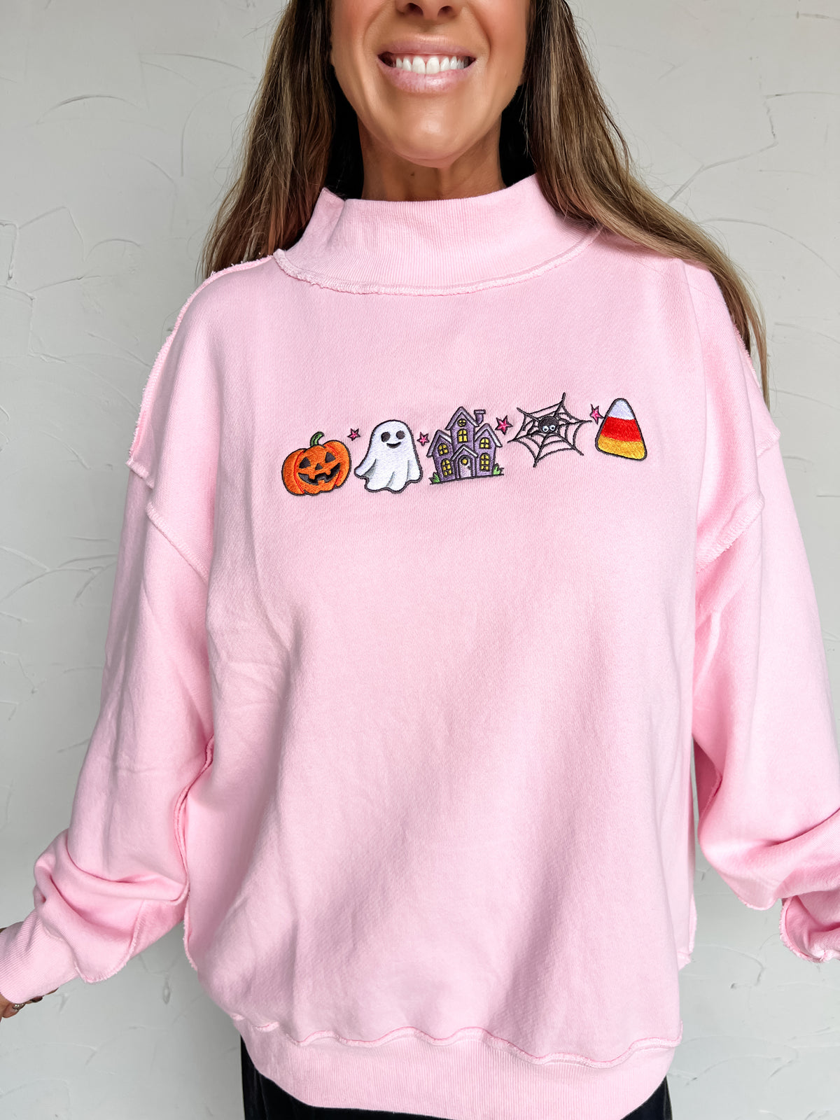 A close-up portrait photograph of a smiling woman with long brown hair posing against a textured off-white wall. She is wearing a light pink long-sleeved sweatshirt made from 100% cotton, featuring a high mock neck collar, drop-shoulder seams with raw-edge reverse stitch detailing, and five embroidered Halloween icons across the chest (a jack-o'-lantern, a friendly ghost, a purple haunted house, a spiderweb, and a piece of candy corn).