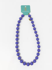 A beaded necklace with large purple beads separated by small gold spacer beads, hanging from a teal card that reads Taylor Shaye Designs, on a white background.