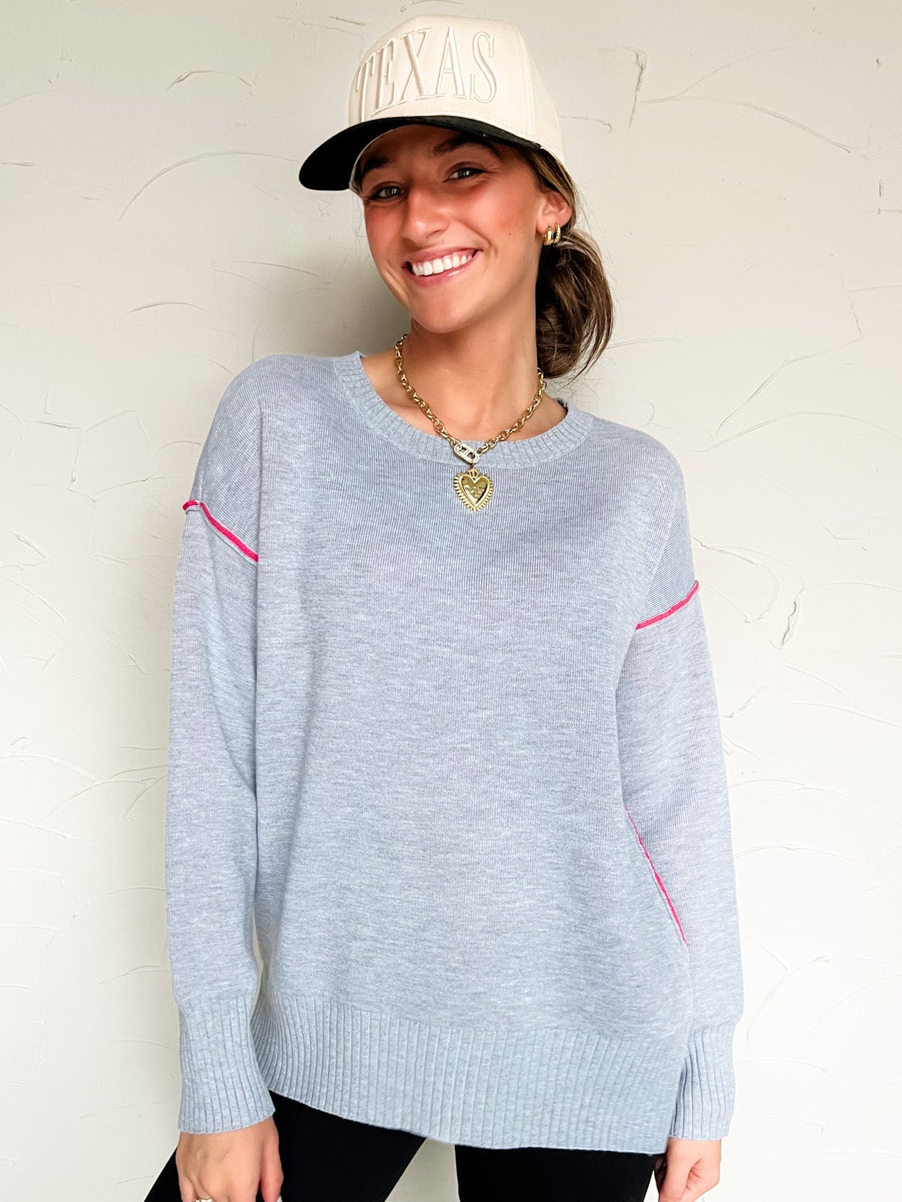 Woman wearing a light grey sweater with pink accents against a white background with a cream cap 