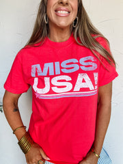 Woman wearing a red 'Miss USA' t-shirt against a white wall
