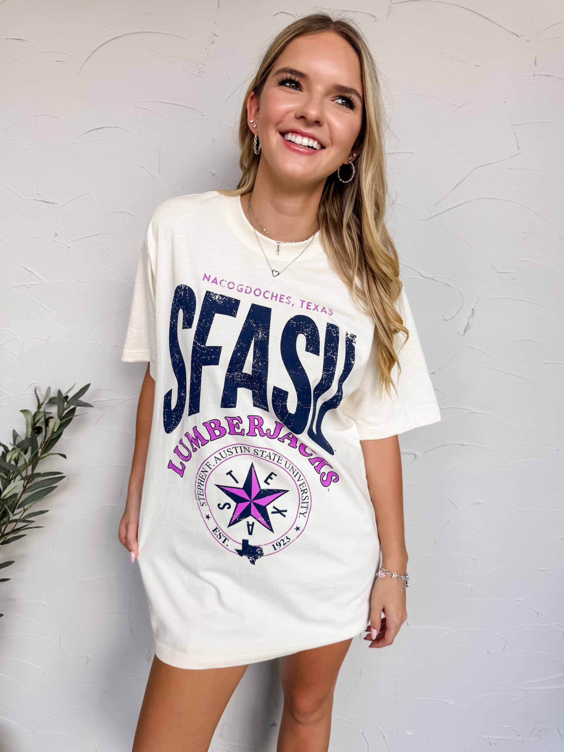 Woman wearing a white t-shirt with 'SFASU Lumberjacks' design against a textured white wall.