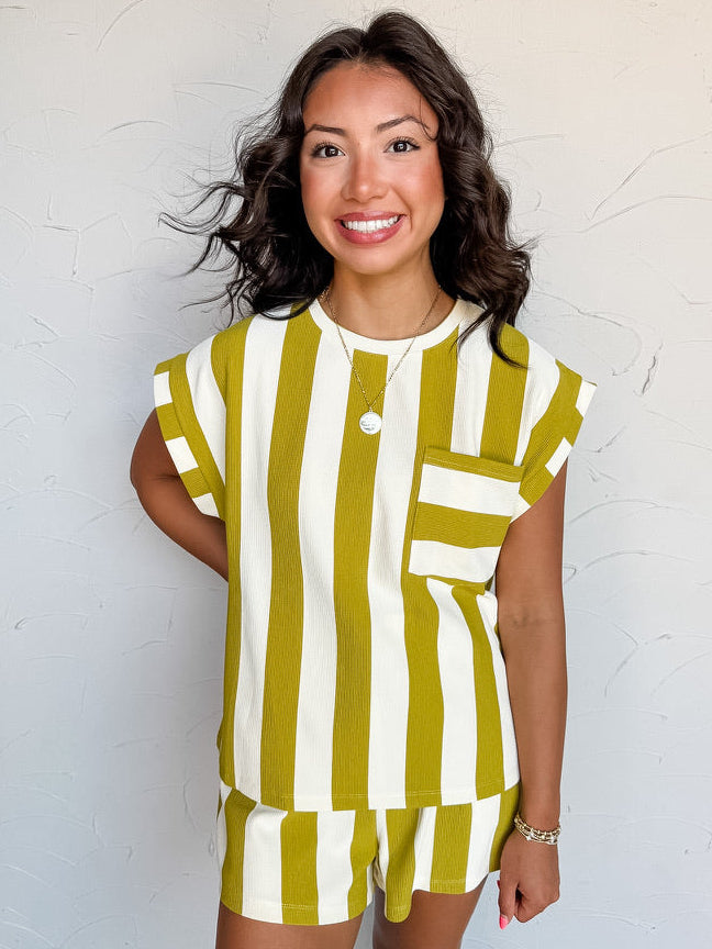 A three-quarter length photograph of a smiling woman with dark wavy hair posing against a textured white wall. She is wearing a short-sleeved top made from a 58% polyester, 30% cotton, 10% rayon, and 2% spandex blend, featuring wide olive green and off-white vertical stripes, a chest pocket, and cuffed cap sleeves, paired with matching striped shorts and styled with a round pendant necklace and gold chain bracelet.