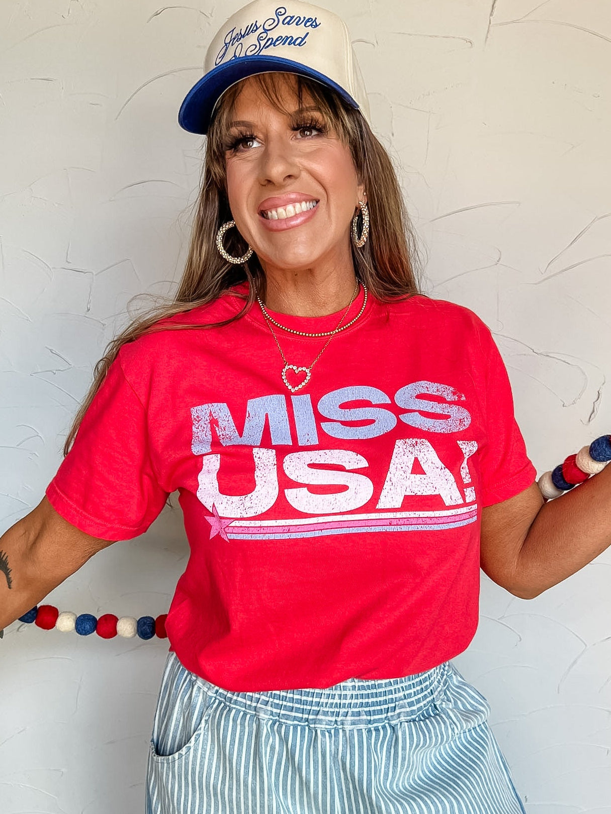 Woman wearing a red 'Miss USA' shirt and blue cap against a white wall.