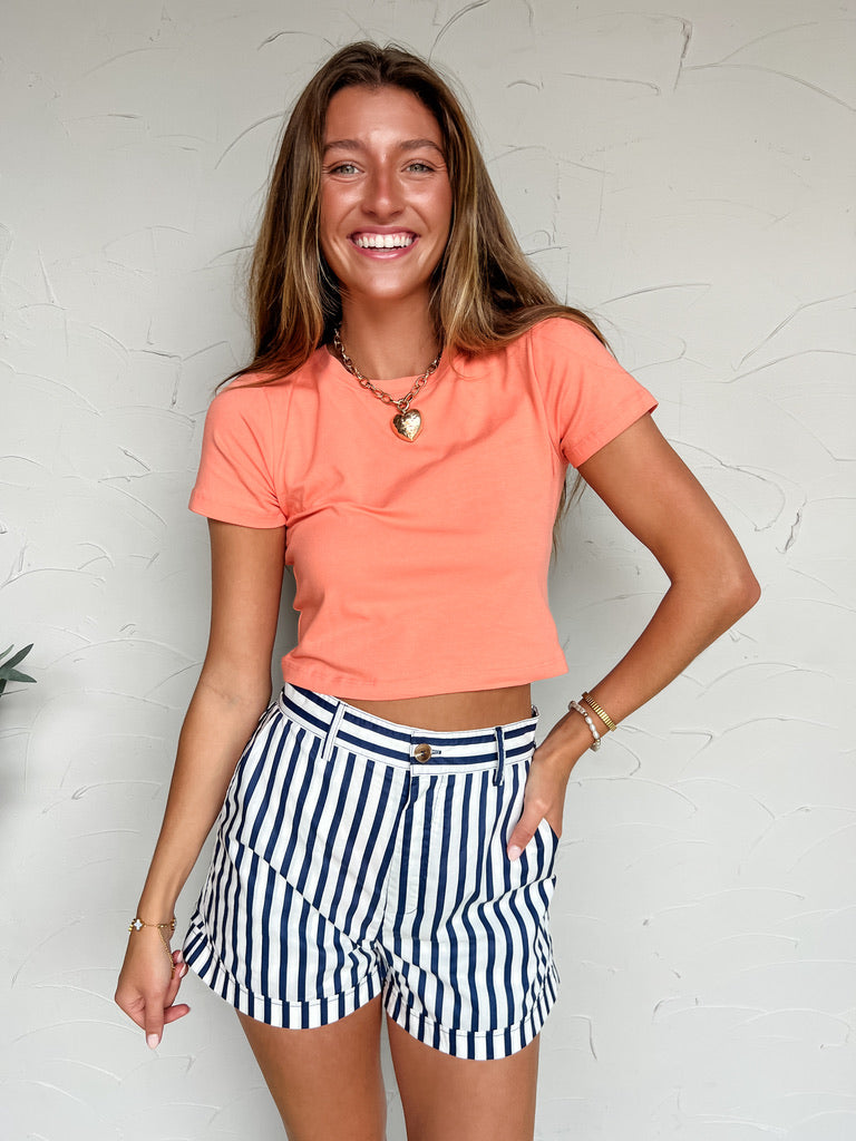 Woman wearing a coral top and striped shorts against a white textured wall