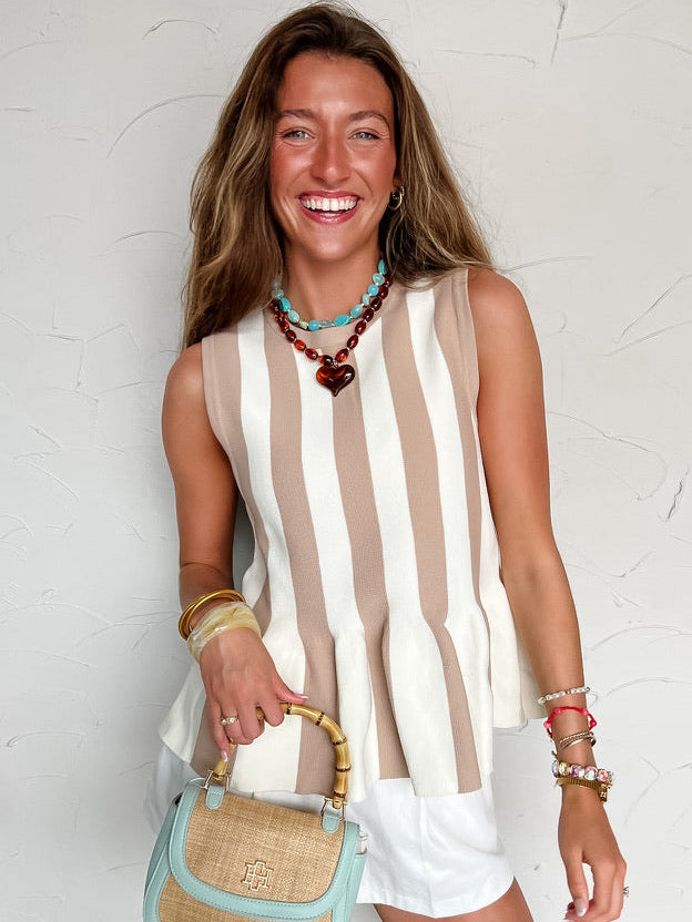 Woman wearing a brown and white striped sleeveless top and white shorts, holding a light blue handbag against a white background.