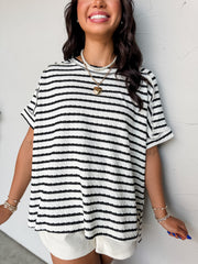 A close-up photograph from the chin down of a smiling brunette woman posing against a textured white wall. She is wearing an oversized crewneck t-shirt in a textured 50% cotton, 43% polyester, and 7% spandex blend, featuring a black and white horizontal stripe pattern with a crinkle-textured finish, dropped shoulders, and short cuffed sleeves.