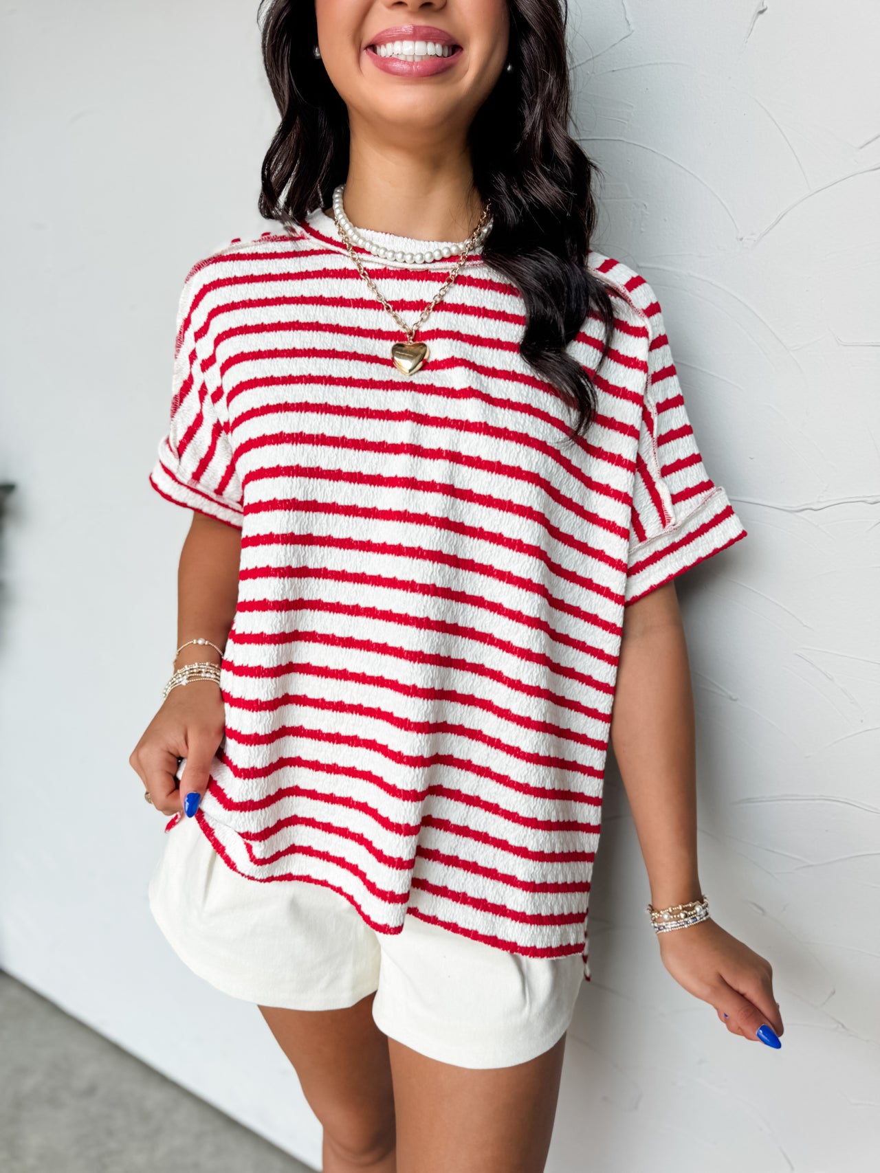 A close-up photograph from the chin down of a smiling brunette woman posing against a textured white wall. She is wearing an oversized crewneck t-shirt in a textured 50% cotton, 43% polyester, and 7% spandex blend, featuring a red and white horizontal stripe pattern with a crinkle-textured finish, dropped shoulders, and short cuffed sleeves.