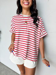 A close-up photograph from the chin down of a smiling brunette woman posing against a textured white wall. She is wearing an oversized crewneck t-shirt in a textured 50% cotton, 43% polyester, and 7% spandex blend, featuring a red and white horizontal stripe pattern with a crinkle-textured finish, dropped shoulders, and short cuffed sleeves.