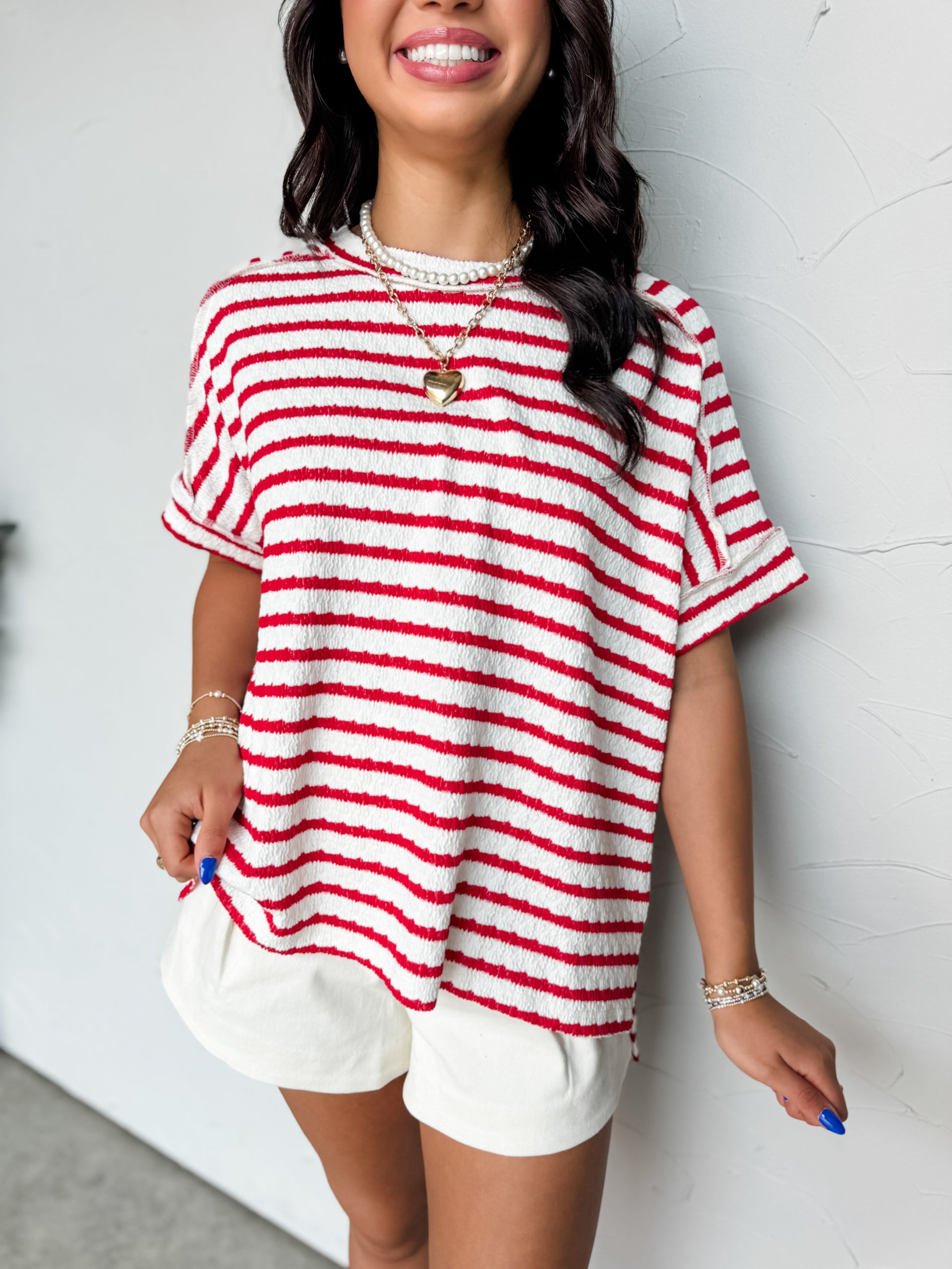 A close-up photograph from the chin down of a smiling brunette woman posing against a textured white wall. She is wearing an oversized crewneck t-shirt in a textured 50% cotton, 43% polyester, and 7% spandex blend, featuring a red and white horizontal stripe pattern with a crinkle-textured finish, dropped shoulders, and short cuffed sleeves.