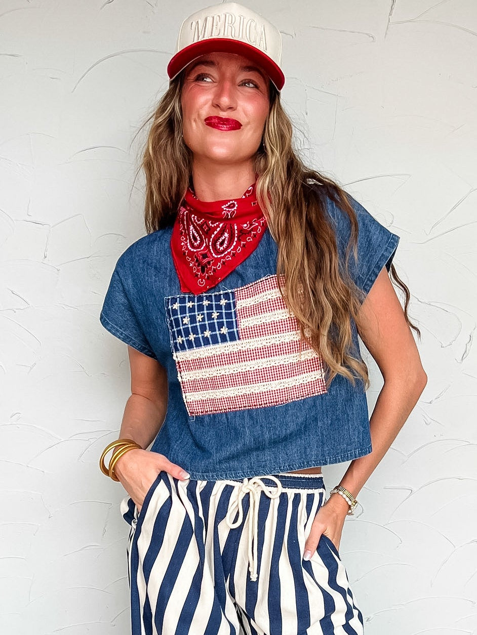 Woman wearing a denim shirt with an American flag design, red bandana, and striped pants against a white wall.