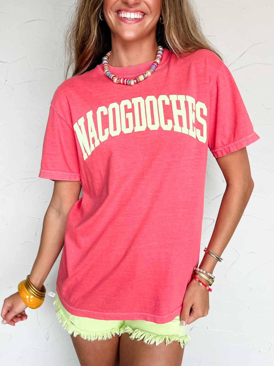 Woman wearing a pink 'Nacogdoches' t-shirt against a white wall