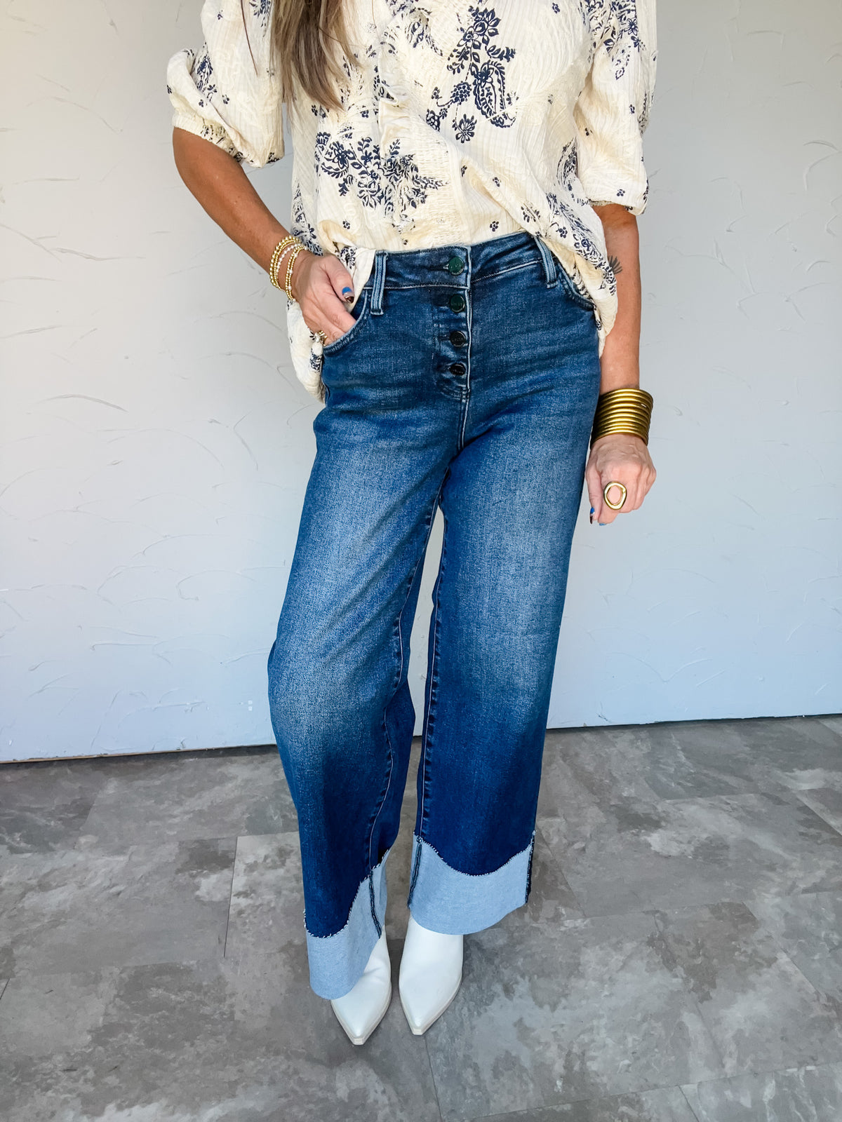 Model wearing Risen Simply Charming Slouchy Wide Leg Cuffed Hem Jeans in dark blue denim, made from 93.5% cotton, 4.5% polyester, and 2% spandex, featuring an exposed button front, front pockets, belt loops, faded thighs, relaxed wide legs, and oversized light blue contrasting cuffs, styled with a cream printed blouse and white pointed-toe ankle boots.