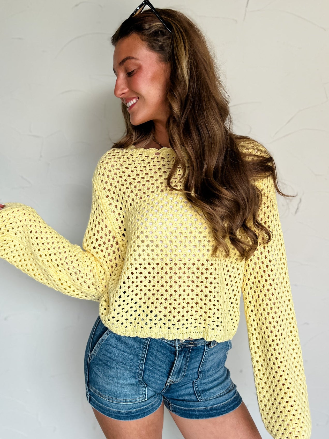 Woman wearing a yellow crochet top and denim shorts against a white wall