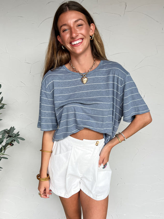 Woman wearing a blue striped shirt and white shorts standing against a white wall.