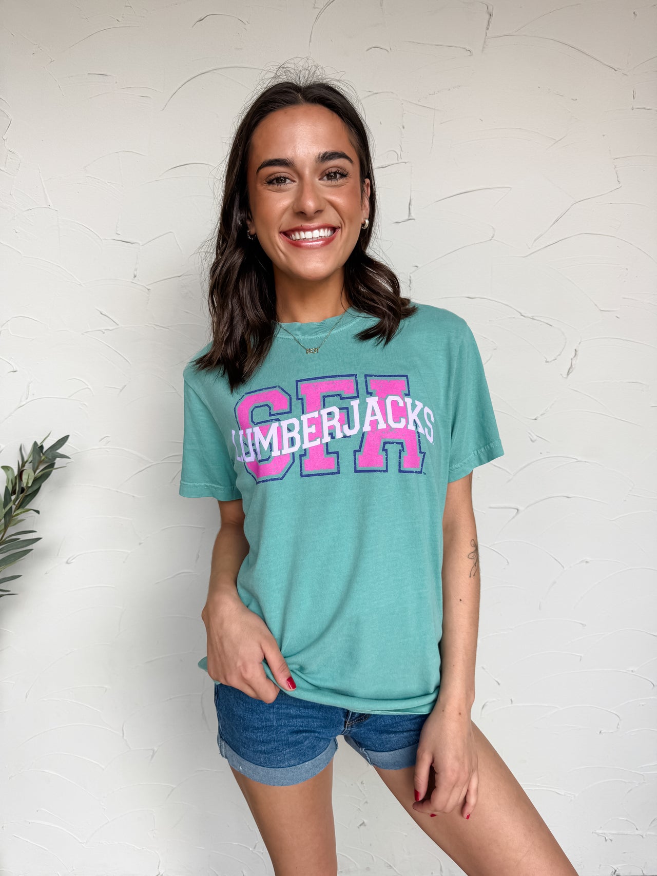 Woman wearing a teal t-shirt with 'SFA' printed on it against a white wall.
