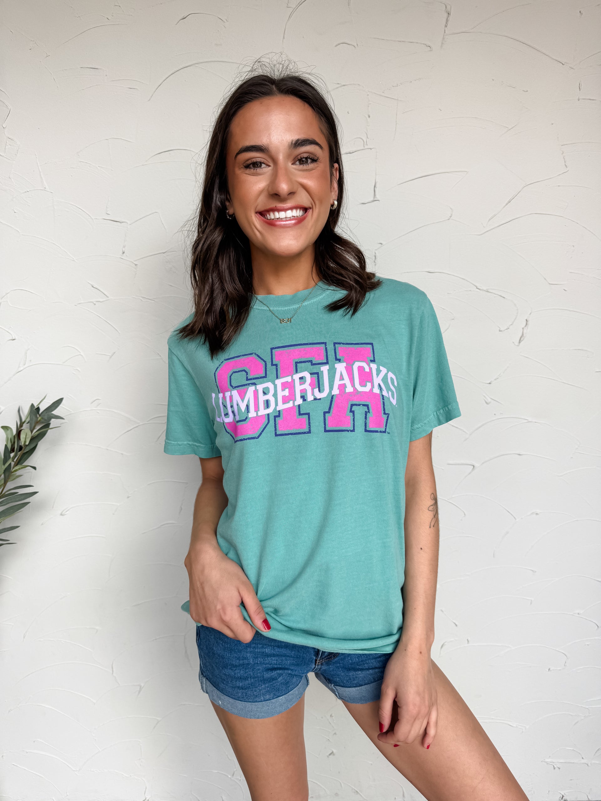 Woman wearing a teal t-shirt with 'SFA' printed on it against a white wall.