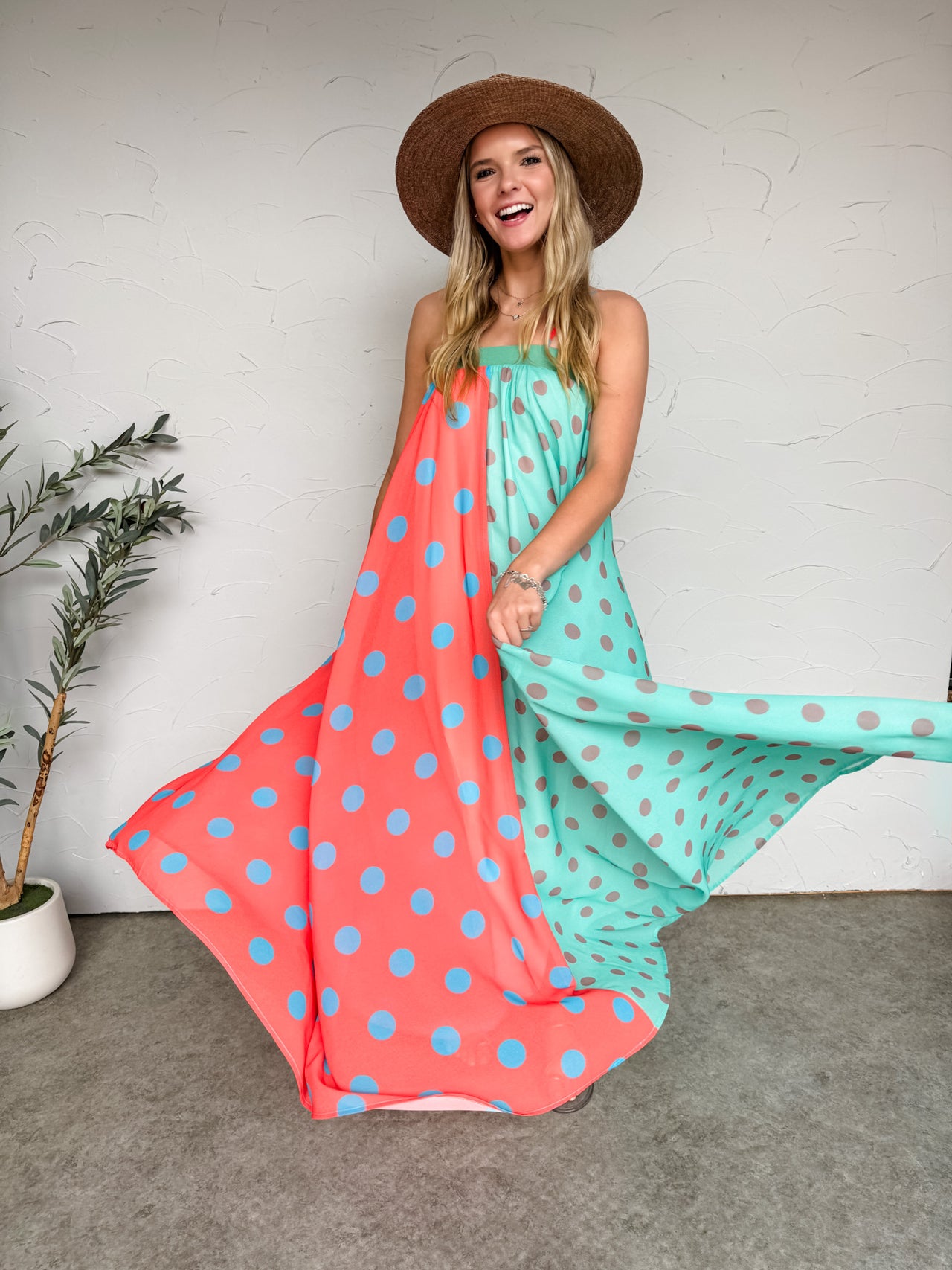 Woman wearing a colorful polka dot dress with a wide-brimmed hat against a white wall.
