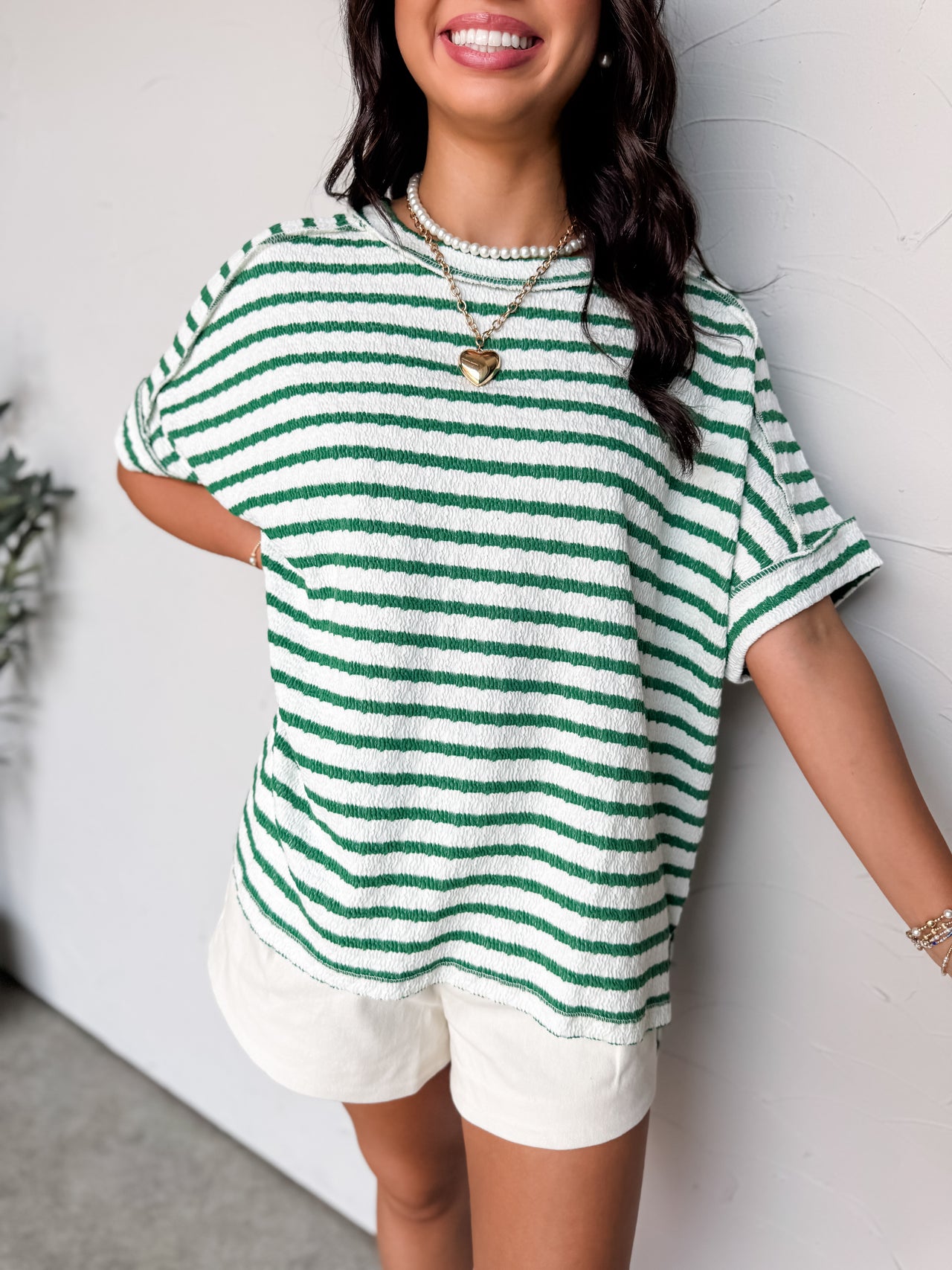 A close-up photograph from the chin down of a smiling brunette woman posing against a textured white wall. She is wearing an oversized crewneck t-shirt in a textured 50% cotton, 43% polyester, and 7% spandex blend, featuring a green and white horizontal stripe pattern with a crinkle-textured finish, dropped shoulders, and short cuffed sleeves.