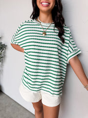 A close-up photograph from the chin down of a smiling brunette woman posing against a textured white wall. She is wearing an oversized crewneck t-shirt in a textured 50% cotton, 43% polyester, and 7% spandex blend, featuring a green and white horizontal stripe pattern with a crinkle-textured finish, dropped shoulders, and short cuffed sleeves.