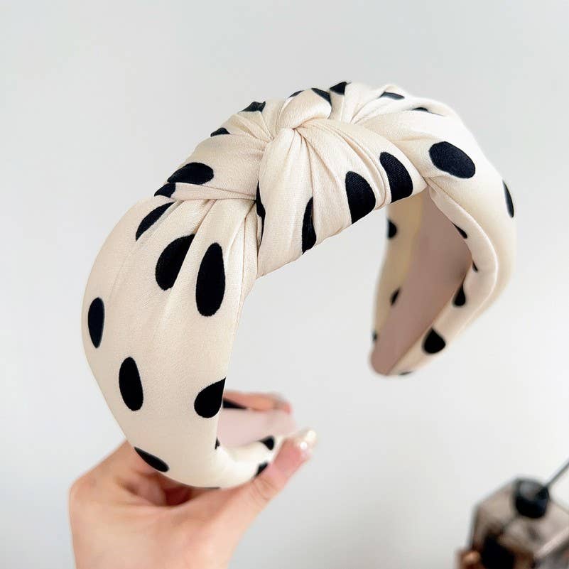 A knotted fabric headband in a warm cream color with black polka dots, held by a hand against a plain light background.