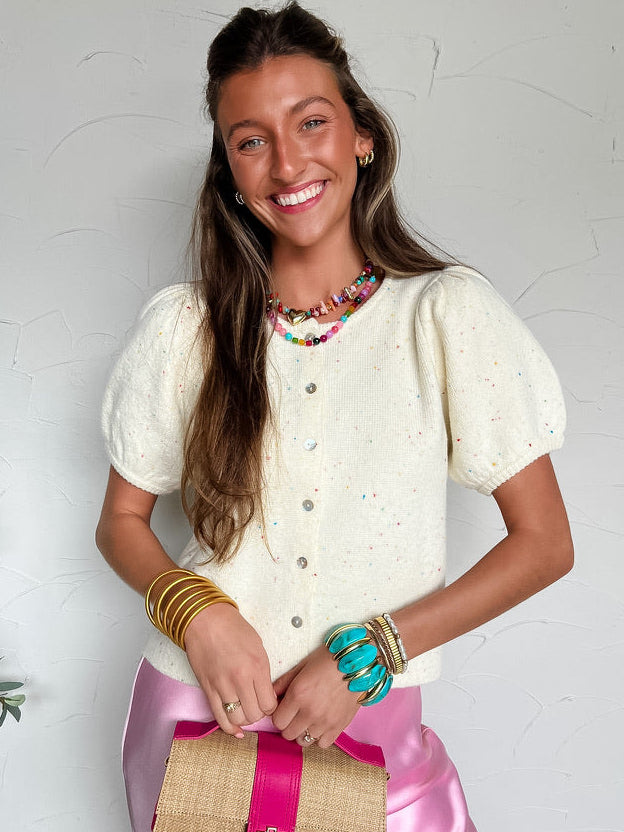 Woman wearing a white knit cardigan with specs of color and pink skirt with a woven bag, standing against a light background.