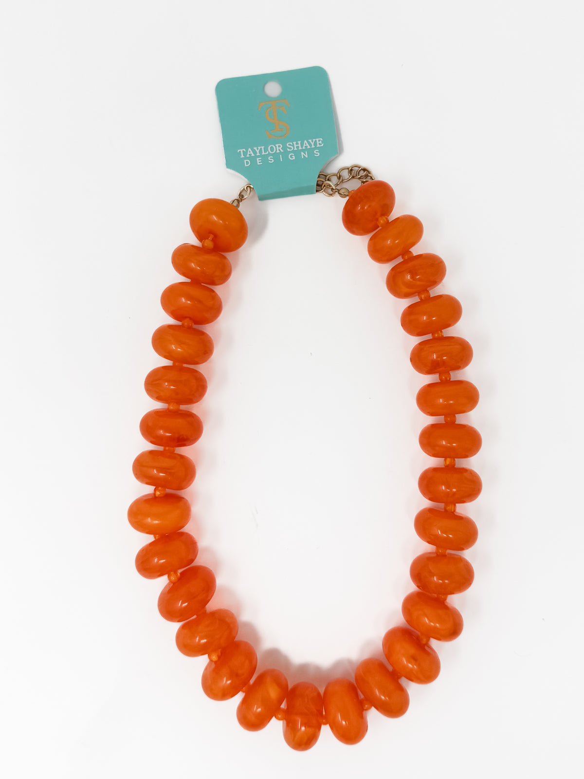 An orange beaded necklace with chunky, rounded disc-like beads, attached to a teal Taylor Shaye Designs brand tag, shown on a plain white background.