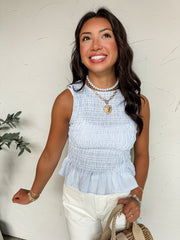 A close-up, front-facing photo of a smiling woman leaning against a textured off-white wall, showcasing a sleeveless, light blue and white pinstriped top. The top features a crew neckline, a tightly smocked bodice that creates an elasticized, textured fit, and a flared ruffle peplum hemline at the waist. She is accessorized with a pearl choker, a chunky gold chain necklace with a heart pendant, large pearl stud earrings, and stacked bracelets. 