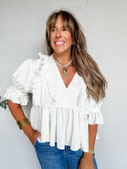 A three-quarter length photograph of a smiling woman with long light brown hair and bangs looking off-camera while posing with one hand in her pocket against a textured white wall. She is wearing a short-sleeved white peplum blouse made from a 65% rayon and 35% nylon blend, featuring a deep V-neckline framed with vertical ruffles, elasticized puff sleeves with ruffled cuffs, and a gathered flared peplum hem, paired with medium-wash blue denim jeans, styled with a layered pearl and chunky gold chain necklace