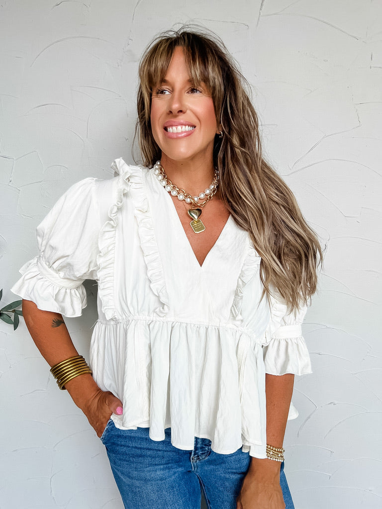 A three-quarter length photograph of a smiling woman with long light brown hair and bangs looking off-camera while posing with one hand in her pocket against a textured white wall. She is wearing a short-sleeved white peplum blouse made from a 65% rayon and 35% nylon blend, featuring a deep V-neckline framed with vertical ruffles, elasticized puff sleeves with ruffled cuffs, and a gathered flared peplum hem, paired with medium-wash blue denim jeans, styled with a layered pearl and chunky gold chain necklace