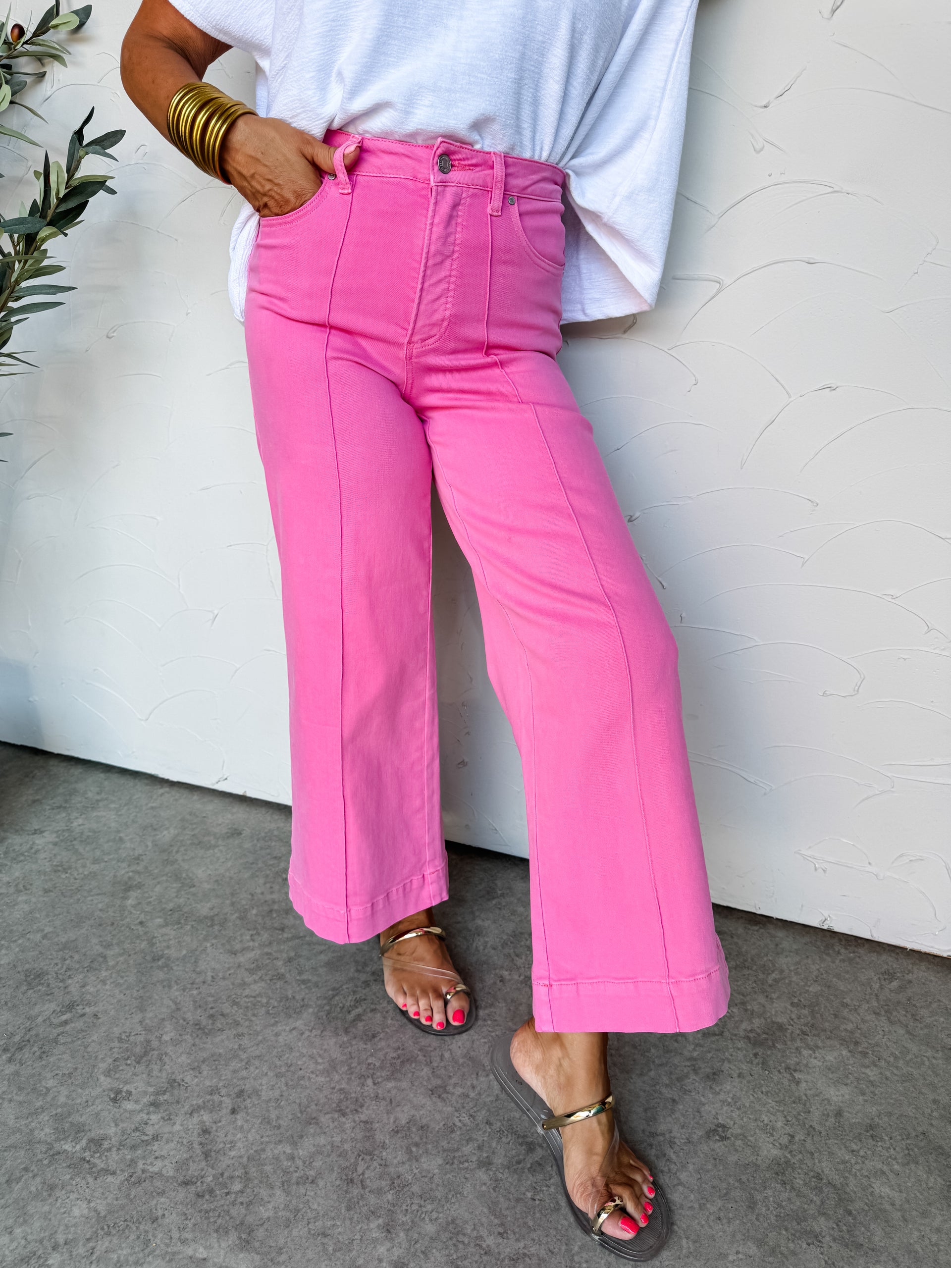 A lower-body view of a person standing against a textured off-white wall, wearing vibrant pink wide-leg pants. The high-waisted trousers are crafted from a stretch denim blend of 91% cotton, 6% polyester, and 3% spandex, featuring a silver button closure, a zip fly, standard pockets, and prominent vertical seams down the center of each leg. They are styled with a tucked-in white textured top and metallic gold flat sandals.