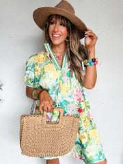 Woman in a green floral dress with a striped collar and straw hat holding a woven bag against a white wall