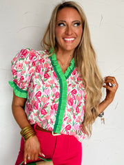 Woman wearing a colorful floral blouse and pink pants, holding a green handbag against a white background.