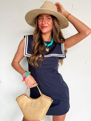 Woman wearing a navy dress with white trim on square collar, straw hat, and holding a straw bag against a white background