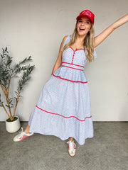 A woman smiling and leaning to the side against a textured off-white wall, wearing a midi-length dress crafted from a 100% cotton seersucker fabrication with a comfortable inner lining. The dress features a light blue and white fine pinstripe base covered in an all-over pink ditsy floral print. It is designed with a sweetheart neckline, a functional button-front placket on the bodice, discrete side seam pockets, and thick shoulder straps that tie at the top. The dress is accented with vibrant red contrast p