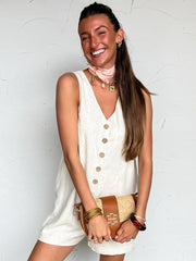Woman in a white sleeveless linen romper standing against a white wall with a plant and woven basket in the background.
