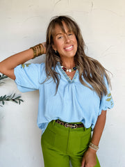 A three-quarter length photograph of a smiling woman with long brown hair and bangs touching her head while posing against a textured off-white wall. She is wearing a light blue short-sleeved blouse made from a textured 95% polyester and 5% spandex fabric blend, featuring embroidered colorful tile-style trim along the split V-neckline and green leafy floral embroidery on the short puff sleeves, front-tucked into olive green trousers with a leopard print belt, styled with a colorful beaded ball necklace, sta