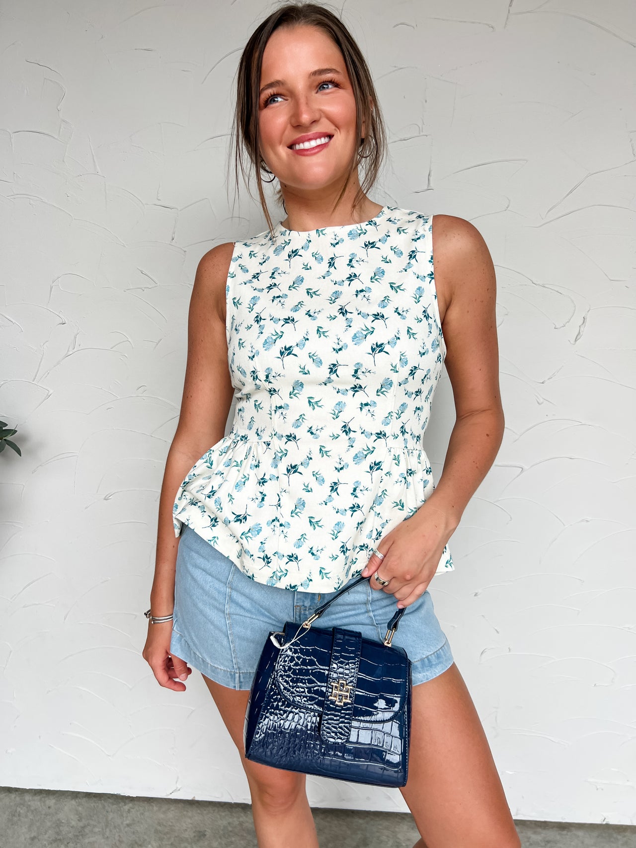 Woman wearing a floral sleeveless top and denim shorts, holding a blue handbag against a white wall.